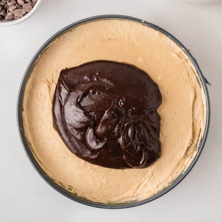A cheesecake in a round pan with a large dollop of glossy chocolate ganache on top, ready to be spread. A bowl of chocolate chips is visible in the background.
