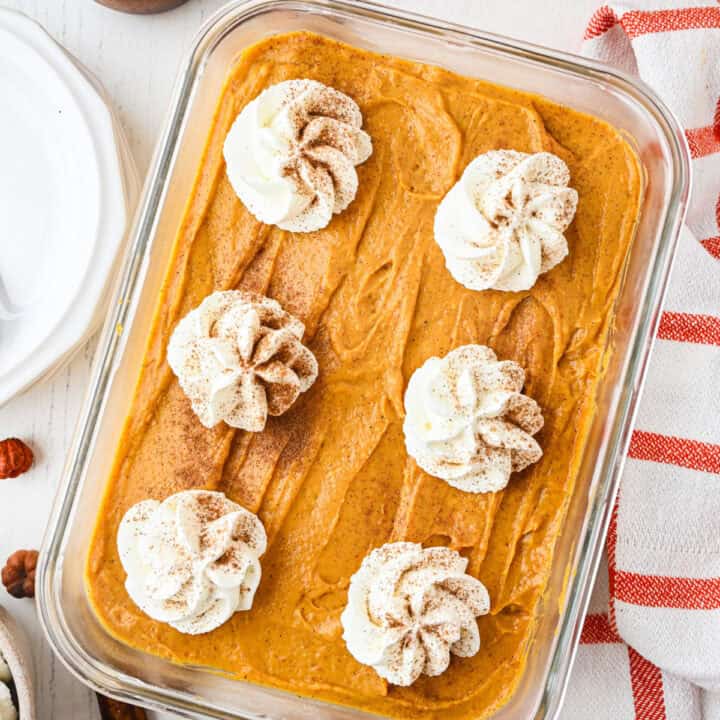 A rectangular glass dish filled with a creamy no-bake pumpkin cheesecake, topped with six swirls of whipped cream and a dusting of cinnamon. A red and white striped towel is nearby.