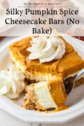 A slice of pumpkin spice cheesecake bar topped with whipped cream sits on a white plate, with a fork holding a bite in the foreground. Text above reads, Silky Pumpkin Spice Cheesecake Bars (No Bake).