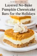 A close-up of a layered no-bake pumpkin cheesecake bar topped with whipped cream and cinnamon, served on a white plate, with another piece blurred in the background. Text reads: Layered No-Bake Pumpkin Cheesecake Bars for the Holidays.