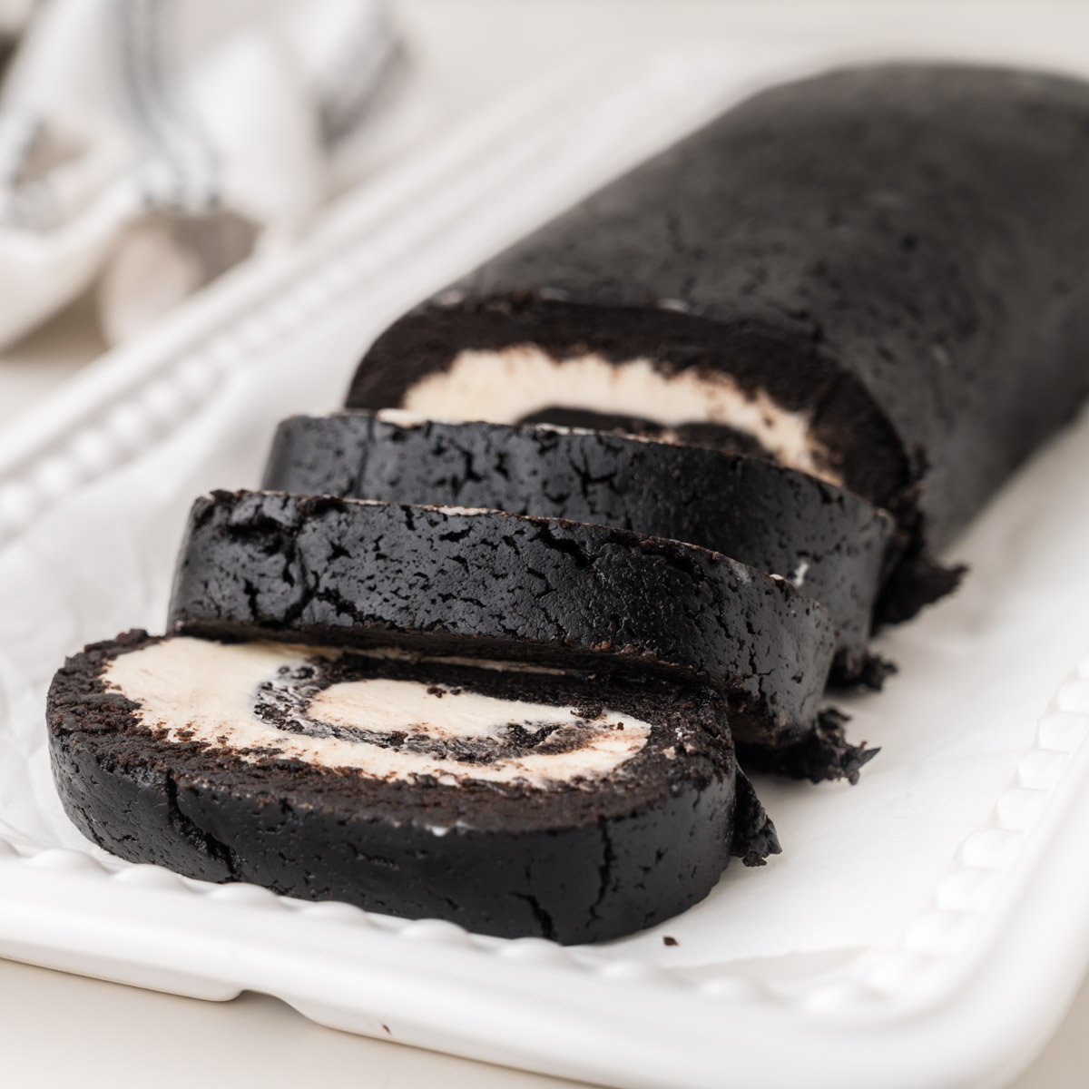 A chocolate oreo cake roll with a white cream filling is sliced and arranged on a white rectangular platter. The roll has a dark, rich appearance and a visible spiral pattern in each slice.