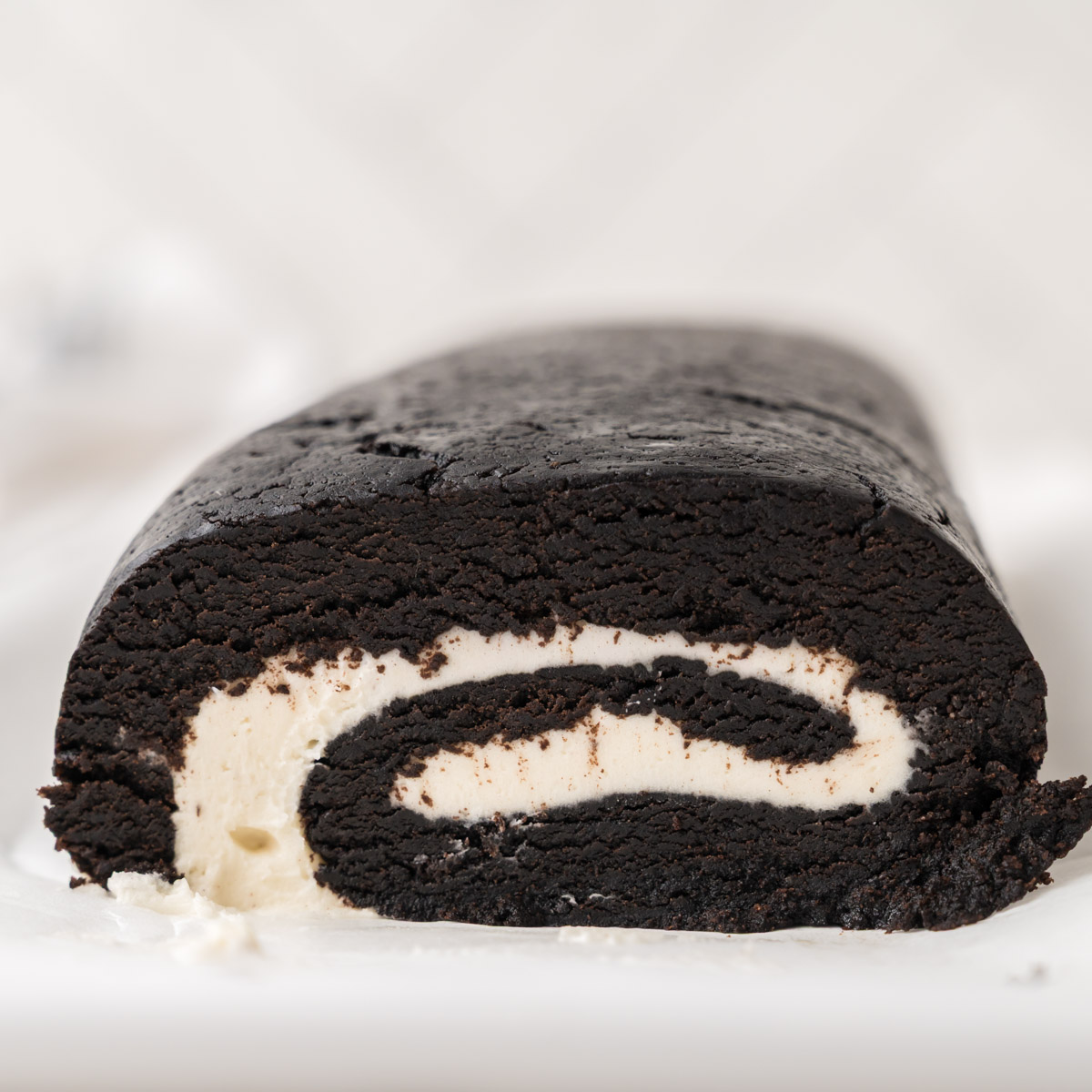 A close-up of a chocolate Swiss roll cake with a creamy white filling, sliced to show the swirl pattern inside. The cake rests on a white surface with a blurred background.
