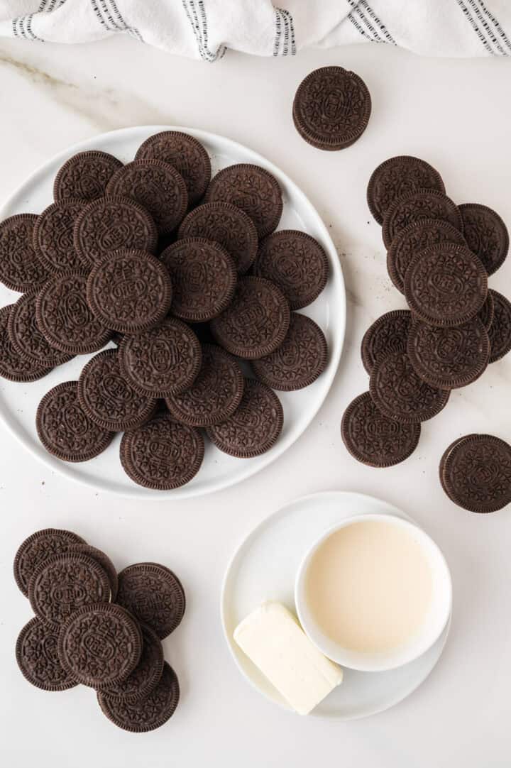 A plate full of chocolate sandwich cookies, more cookies stacked nearby, and a cup of milk with a slab of butter on a white saucer, all on a white surface with a striped towel at the top.