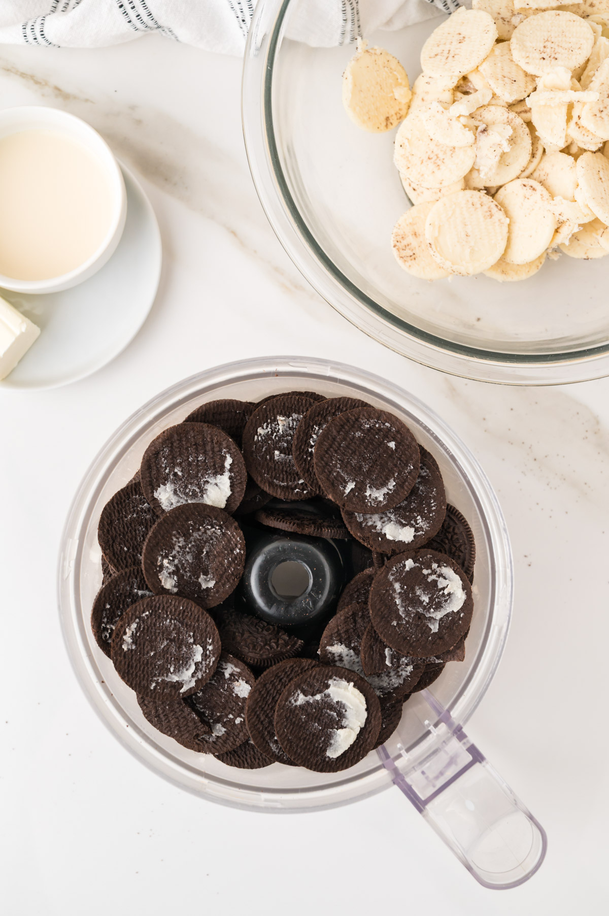A food processor filled with chocolate sandwich cookies, partially separated, sits on a white surface near a clear bowl of vanilla wafer cookies, a small plate of butter, and a cup of milk.