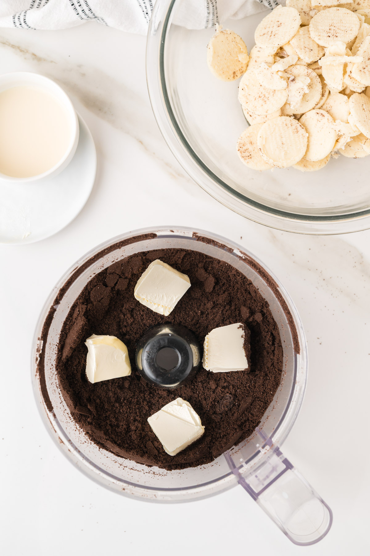 Overhead view of a food processor with crushed chocolate cookies and butter, a bowl of sliced bananas, and a cup of milk on a white countertop.