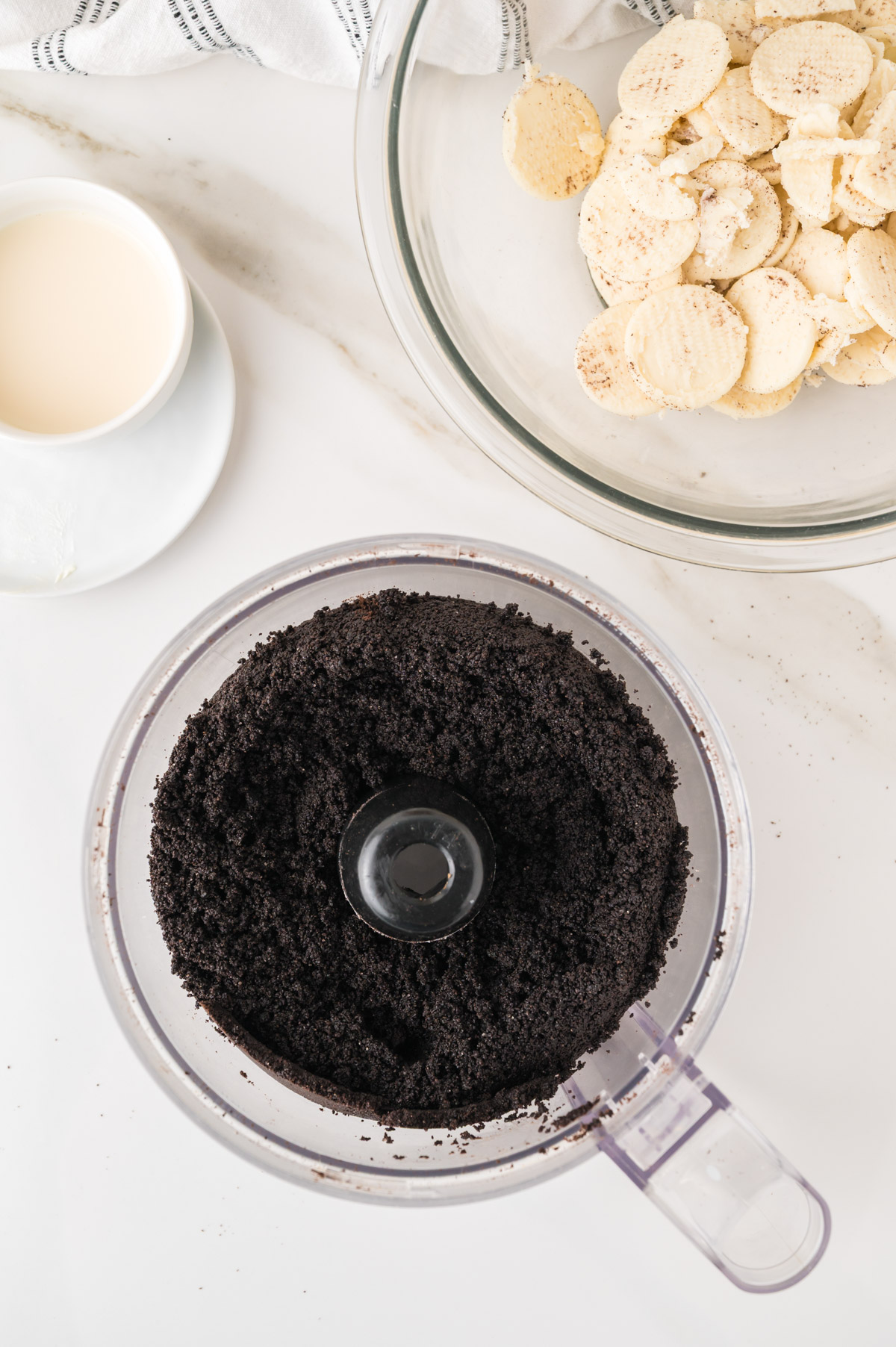 A food processor filled with crushed chocolate cookies sits on a white countertop next to a large glass bowl of sliced bananas and a small cup of milk.