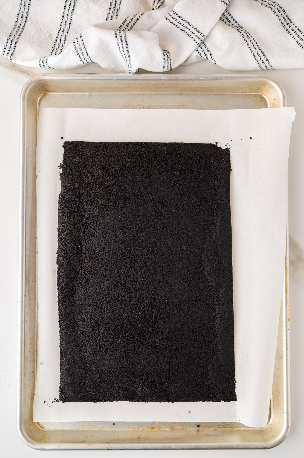 A rectangular sheet of dark, crumbly cookie crust on parchment paper rests on a baking tray, with a striped kitchen towel partially visible at the top.