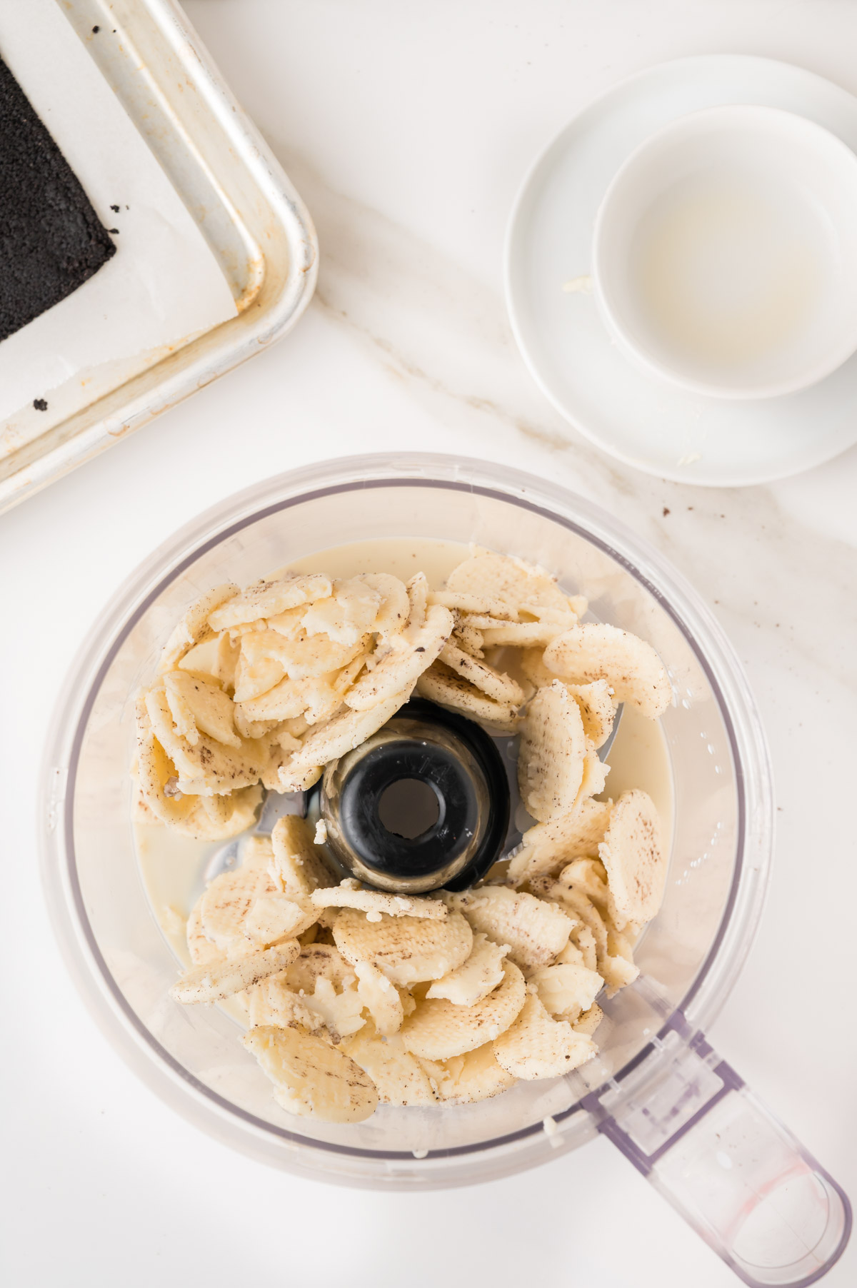 Overhead view of a food processor filled with broken cookie pieces, next to a baking tray with a dark rectangular baked item and a white plate with a small bowl holding some liquid, all on a white countertop.