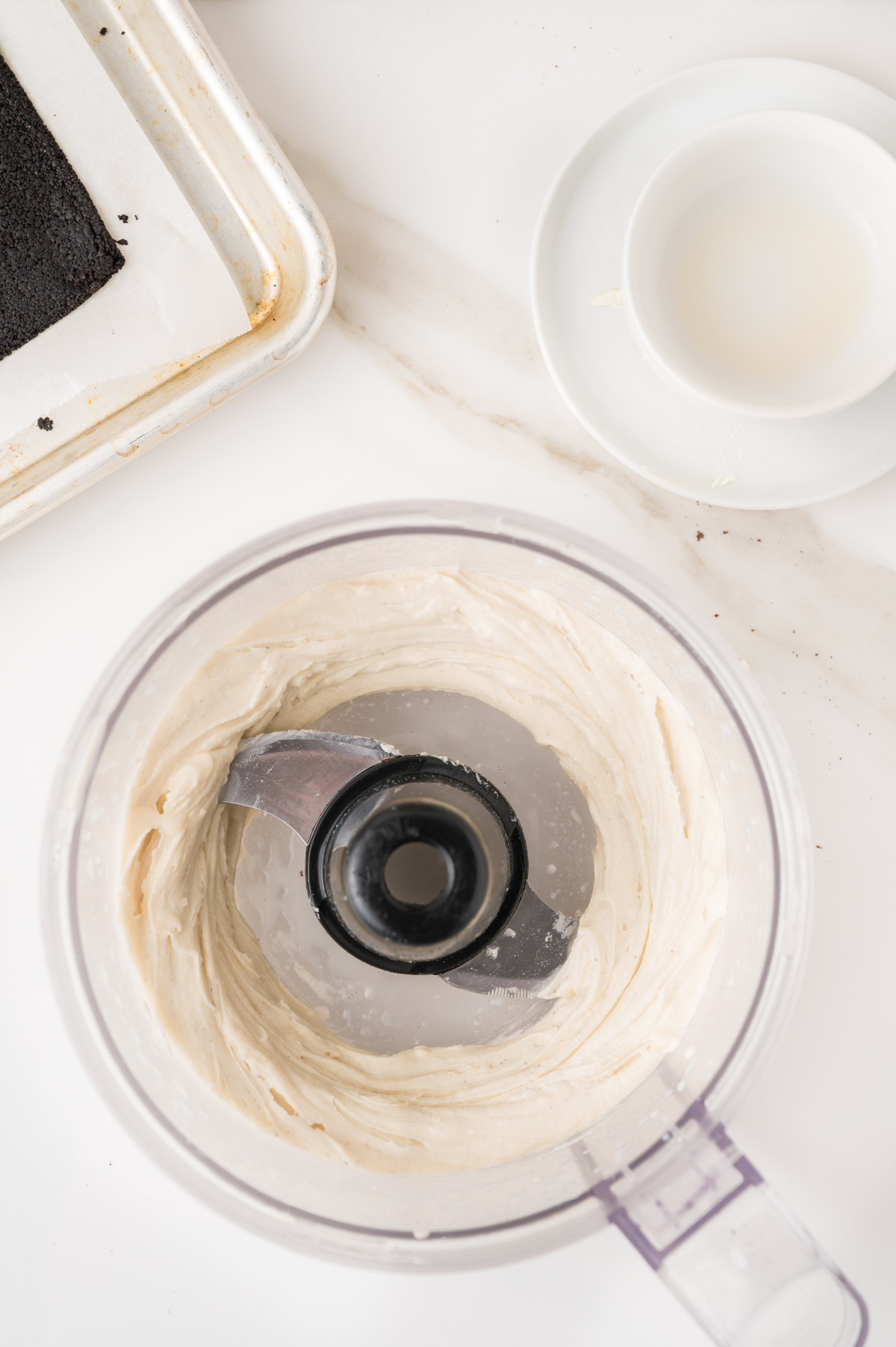 A food processor with creamy white frosting inside is on a white surface. Next to it is a small dish and a baking tray with a piece of chocolate cake.