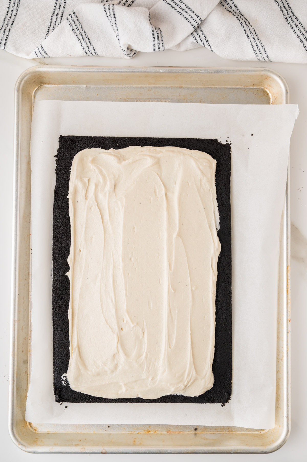 A baking sheet with parchment paper holds a rectangular layer of dark cookie crust topped with a smooth, creamy white filling. A striped kitchen towel is partially visible above the pan.