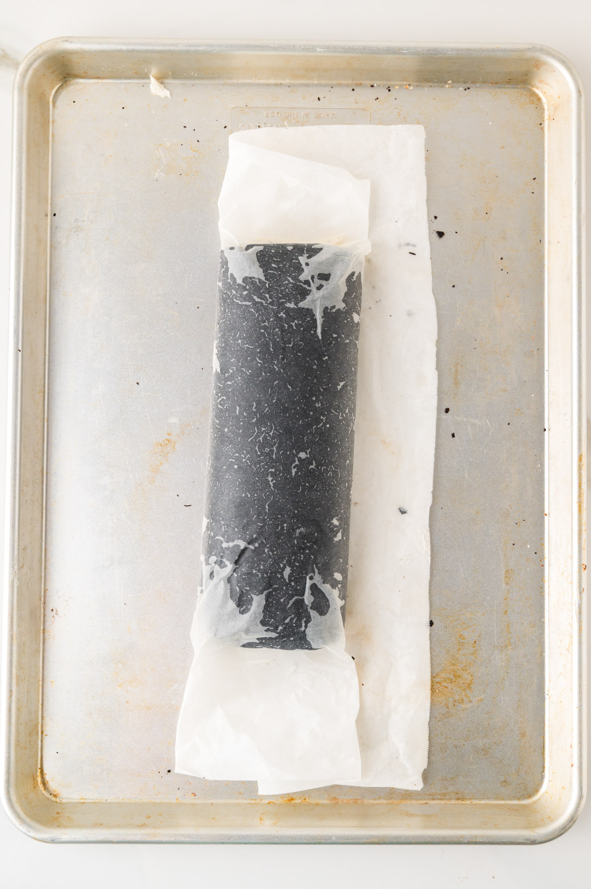 A rolled log of dark dough wrapped in parchment paper sits on a metal baking sheet with light stains.