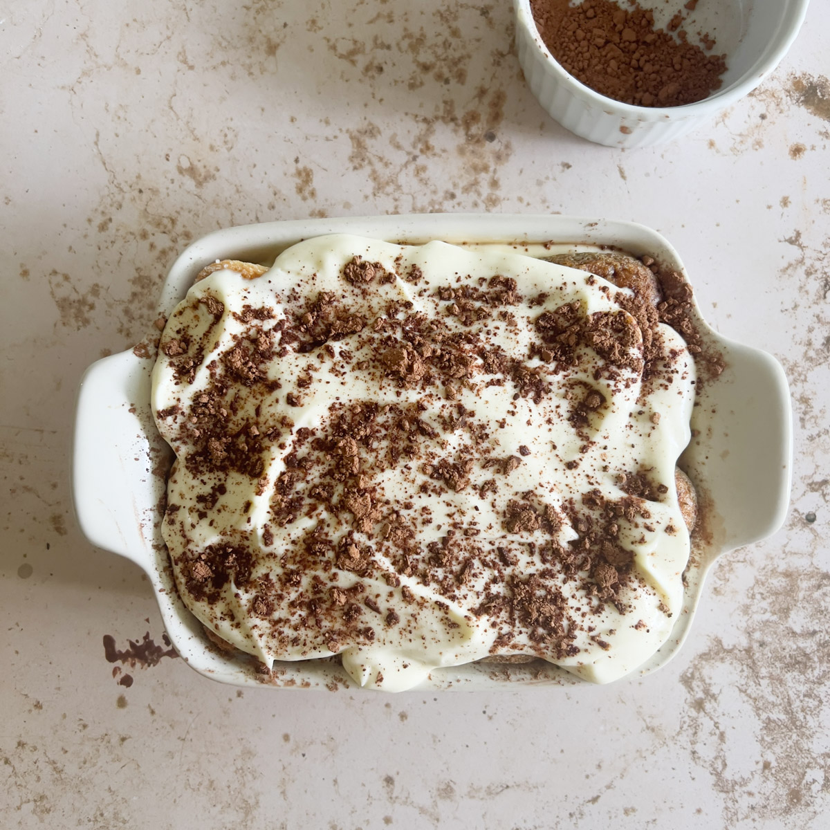 A white ceramic dish filled with tiramisu, topped with creamy mascarpone and sprinkled with cocoa powder. In the background, a small white bowl holds extra cocoa powder on a light marbled surface.