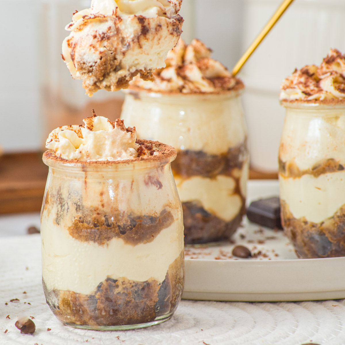 Three glass jars filled with layers of creamy tiramisu and coffee-soaked cake, topped with whipped cream and cocoa powder. A spoon lifts a portion from one jar, showcasing the desserts rich, layered texture.