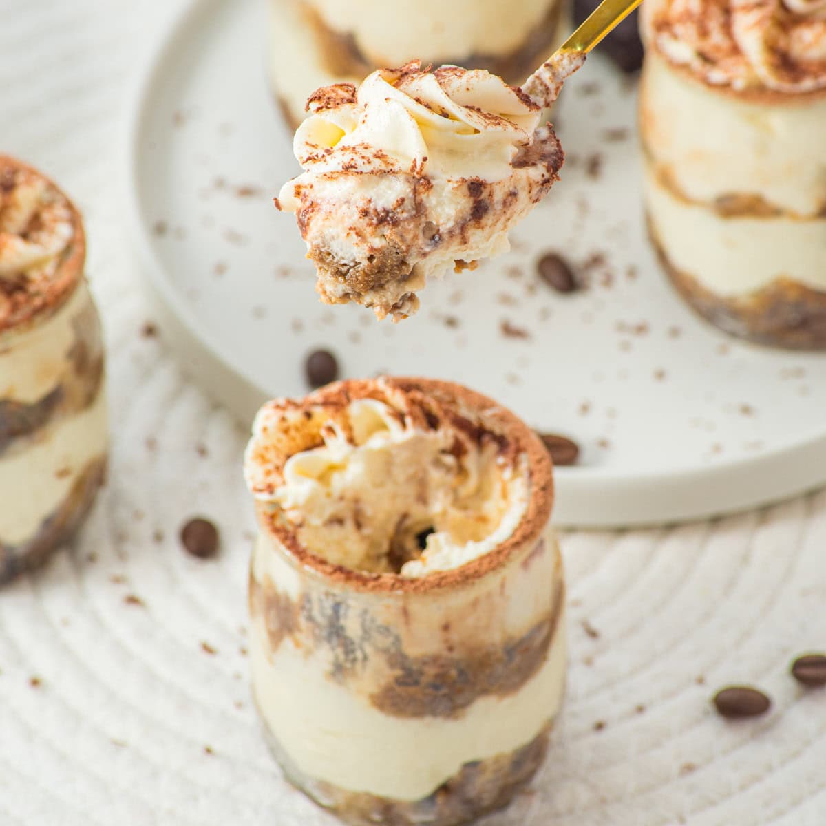 A close-up of a spoon holding a bite of creamy tiramisu, with jars of tiramisu and scattered coffee beans on a white textured surface in the background.
