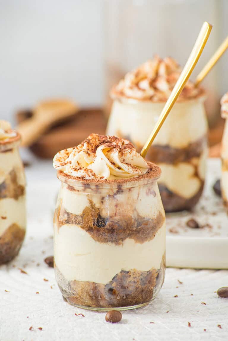 A small glass jar filled with layers of creamy tiramisu and cocoa-dusted whipped topping, with a gold spoon inside. More jars of tiramisu are blurred in the background.