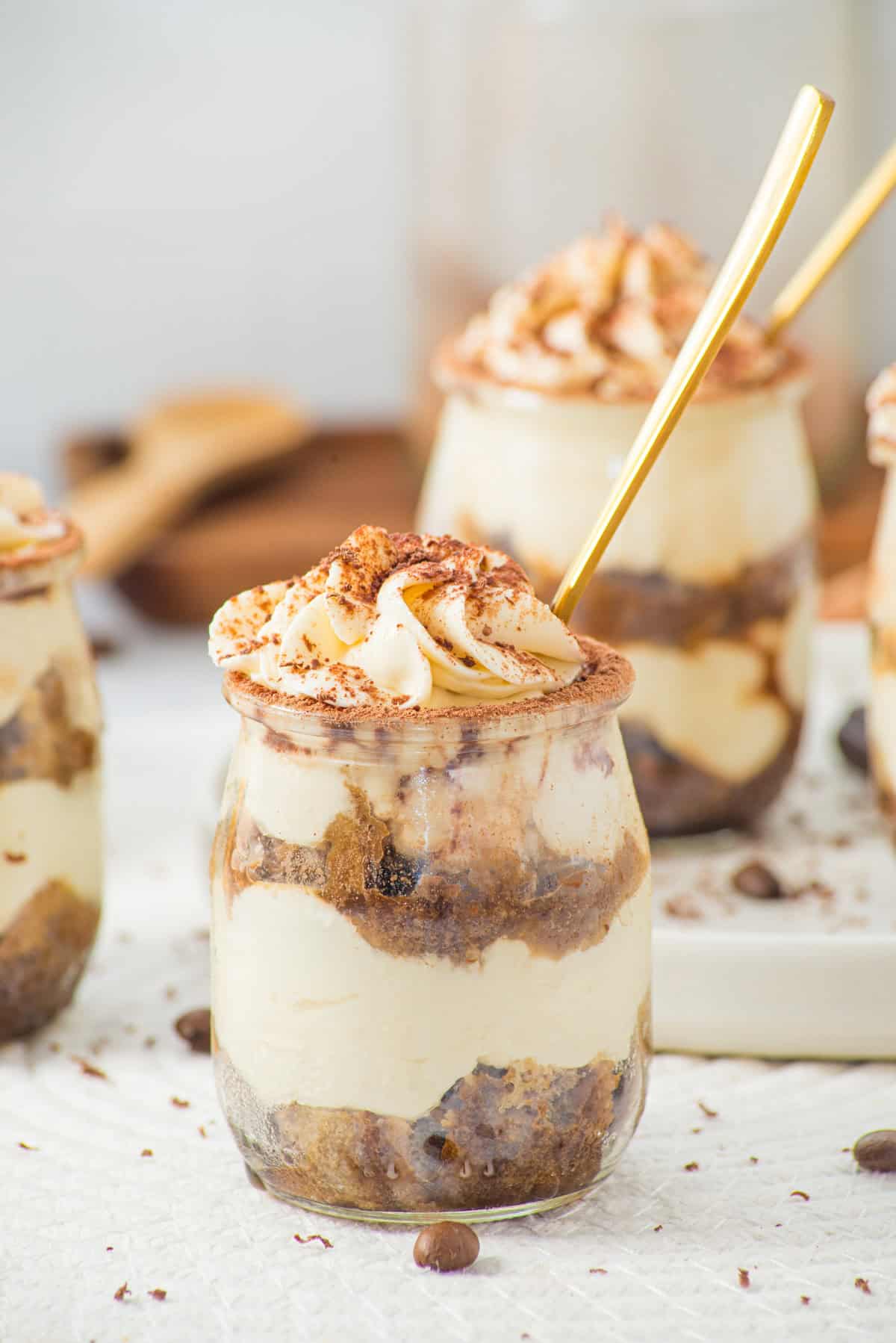 A small glass jar filled with layers of creamy tiramisu and cocoa-dusted whipped topping, with a gold spoon inside. More jars of tiramisu are blurred in the background.