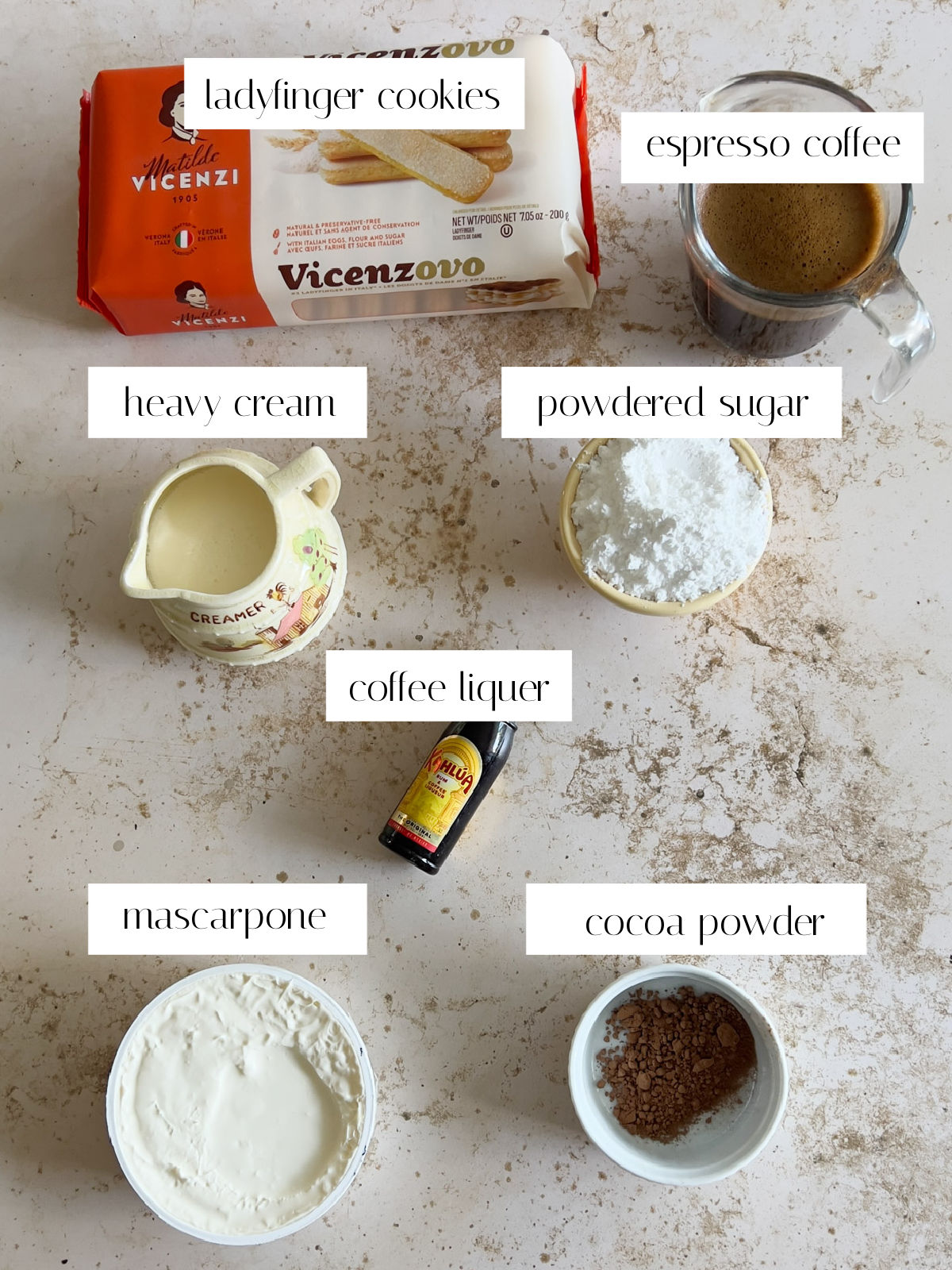 Top-down view of ingredients for tiramisu, including ladyfinger cookies, espresso coffee, heavy cream, powdered sugar, coffee liqueur, mascarpone cheese, and cocoa powder, each labeled on a marble surface.